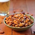 A bowl of spicy nuts near a glass of beer on a wooden board.