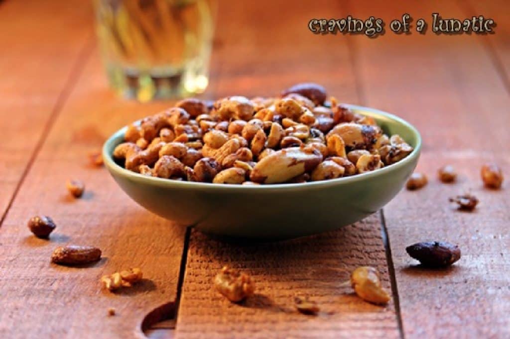 A bowl of spicy nuts on a wooden board with a glass of beer in the background.
