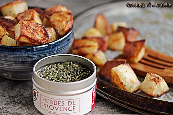 Perfect Roast Potatoes in a bowl and on a pan with a wooden spatula on it. A tin of herbs is in the foreground. 