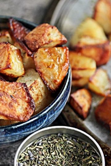 Roast potatoes in a blue bowl with a jar of herbs nearby and more potatoes spilled on counter.