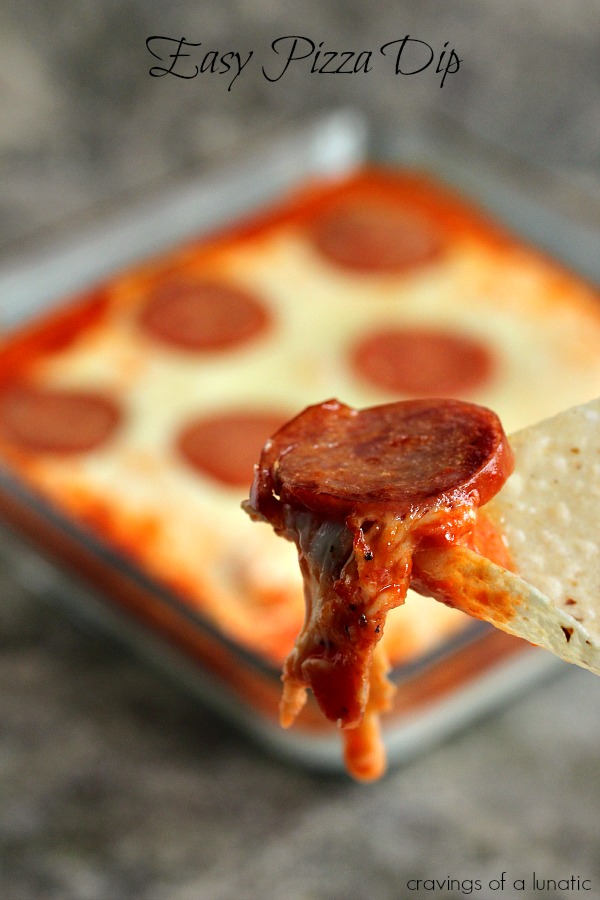 A close up of pizza dip on a chip with a bowl of the remaining dip in the background.
