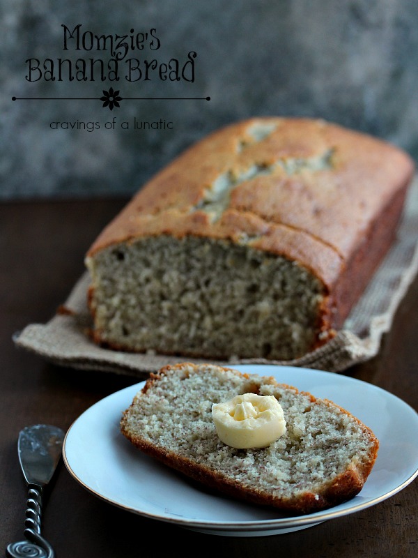 A slice of banana Bread on a white plate with a pat of butter on it. The rest of the loaf of banana bread is in the background. 