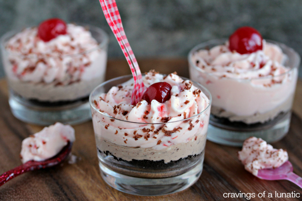 No bake cherry and chocolate cheesecakes layered in tiny glasses with cherry on top and spoon inside a glass and on a wood board. 