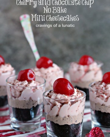 No bake cherry and chocolate chip cheesecake layered in shot glasses with a cherry on top and spoon in some glasses. Shot glasses are sitting on a red and white napkin with a dark background.