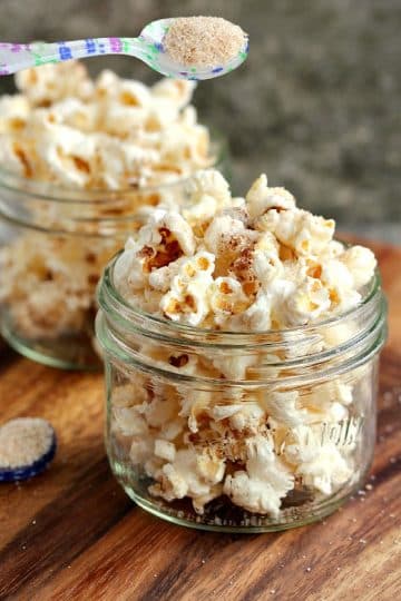 Cinnamon sugar popcorn served in two small mason jars with a spoonful of cinnamon sugar being sprinkled over top.