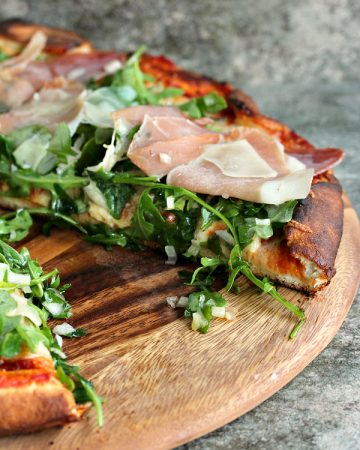 Pizza topped with sauce, cheese, arugula and prosciutto served on a wood board.