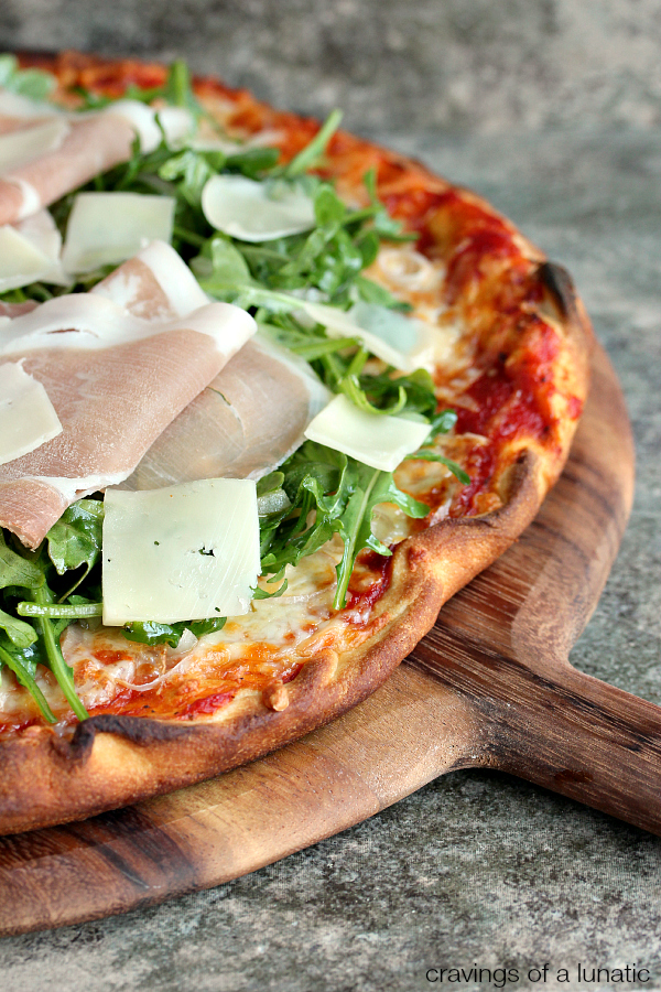 Pizza topped with sauce, cheese, arugula and prosciutto served on a wood board. 