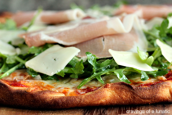 Pizza topped with sauce, cheese, arugula and prosciutto served on a wood board. 