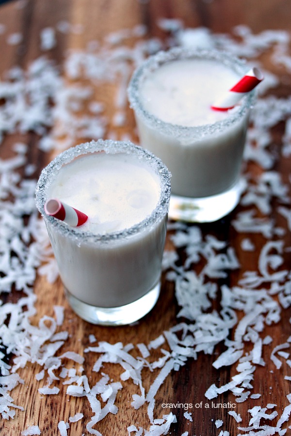 Mini Coconut Milkshakes served in shot glasses with decorative rims and tiny straws inside. Grated coconut is scattered on a wood tray around the milkshakes.