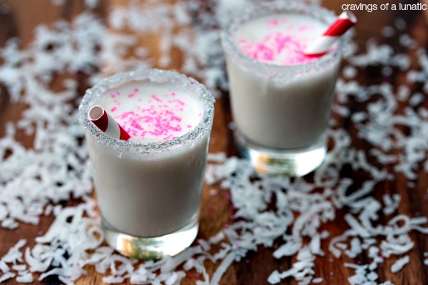 Mini Coconut Milkshakes served in shot glasses with decorative rims and tiny straws inside. Grated coconut is scattered on a wood tray around the milkshakes.