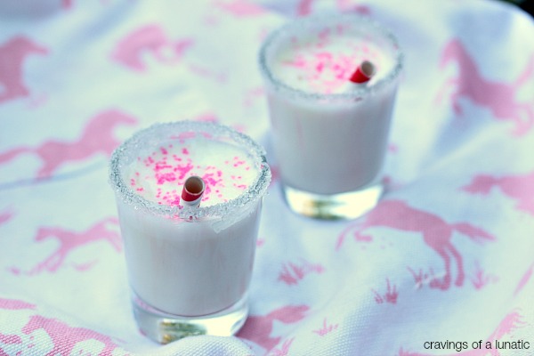 Mini coconut milkshakes served in shot glasses with decorative rims and places on a white and pink cloth napkin with horses on it.