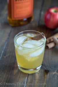 Bourbon Apple Cider in a glass with ice and a cinnamon stick. Glass is on a wood board with ingredients in the background.