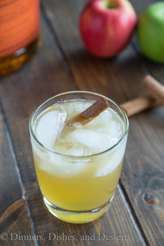 Bourbon Apple Cider in a glass with ice and a cinnamon stick. Glass is on a wood board with ingredients in the background. 