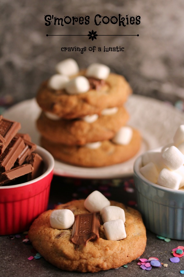 Smores Cookies image, cookies on a white plate with one cookie on the counter in front of it. A small red bowl is holding chocolate and a small blue bowl is holding mini marshmallows. 