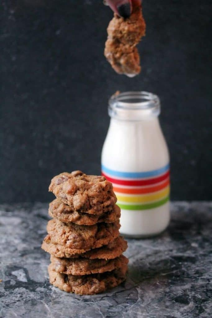 A stack of chocolate chip pistachios cookies on a counter with a glass of milk in the background with a cookie being dunked into it. 
