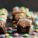 M&M brownies topped with frosting and M&Ms on a counter with lots of pastel m&ms on it.