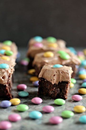 M&M brownies topped with frosting and M&Ms on a counter with lots of pastel m&ms on it.