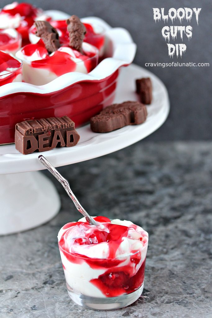 Bloody guts cherry dip served in a pie plate with walking dead chocolates, pie dish is sitting on a white cake plate and there is a small glass serving bowl filled with the dip and a spoon in the foreground. 