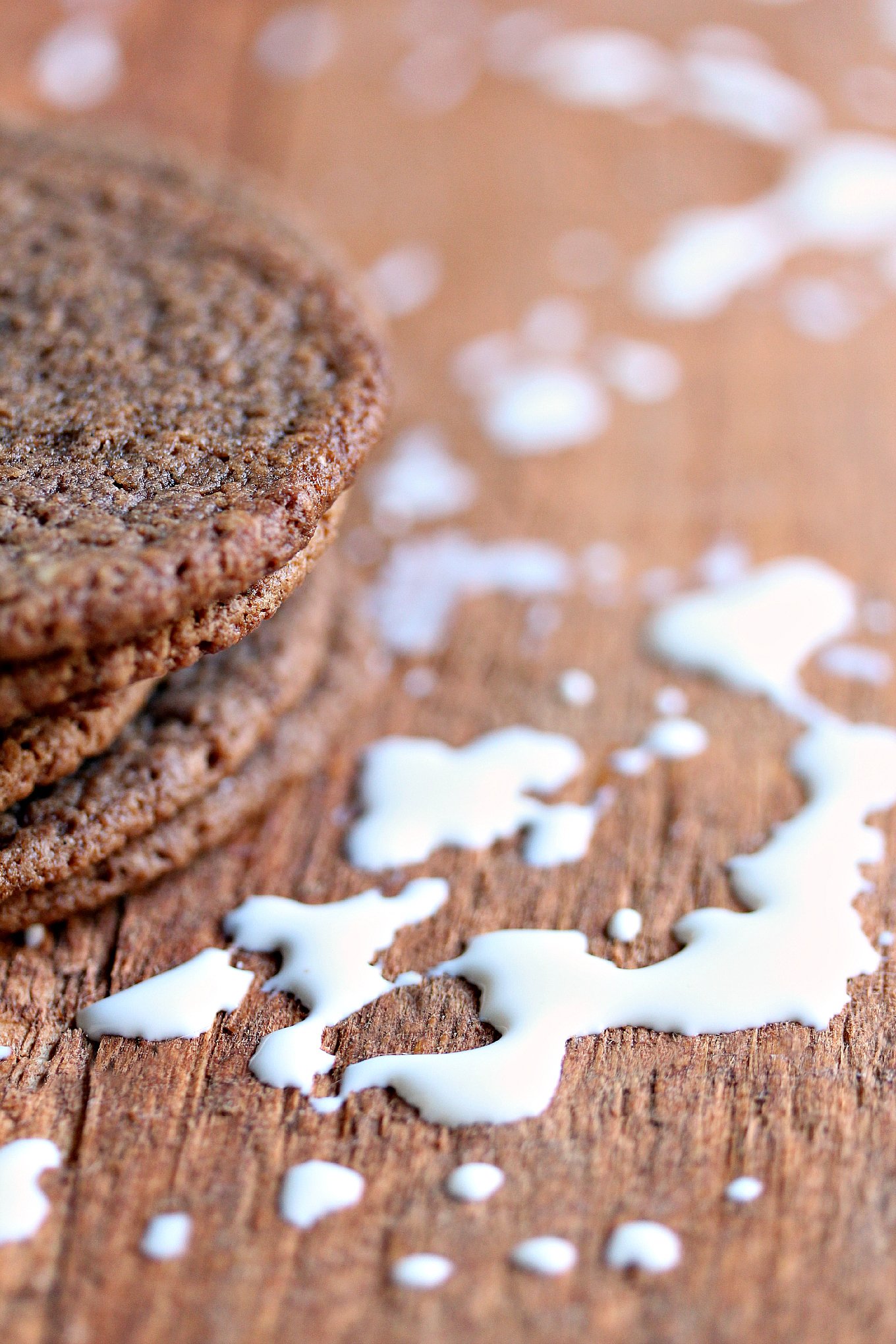 Carol's chewy chocolate cookies stacked on a wood surface with milk spilled all over it. 