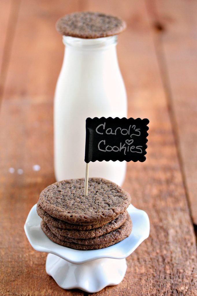 Chewy chocolate cookies stacked on a mini cupcake stand with a tiny chalkboard stuck in them that has the recipe name written on it, with a bottle of milk in the background.