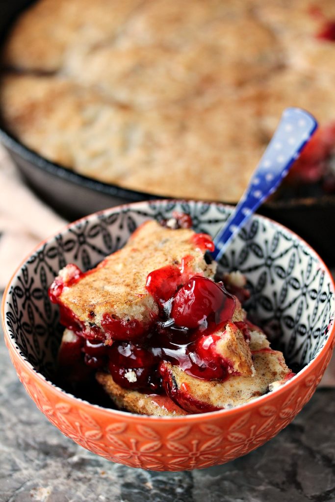 Cherry cobbler served in a small orange bowl with a blue and white polka dot spoon. Cast iron skillet filled with cobbler can be seen in the background.
