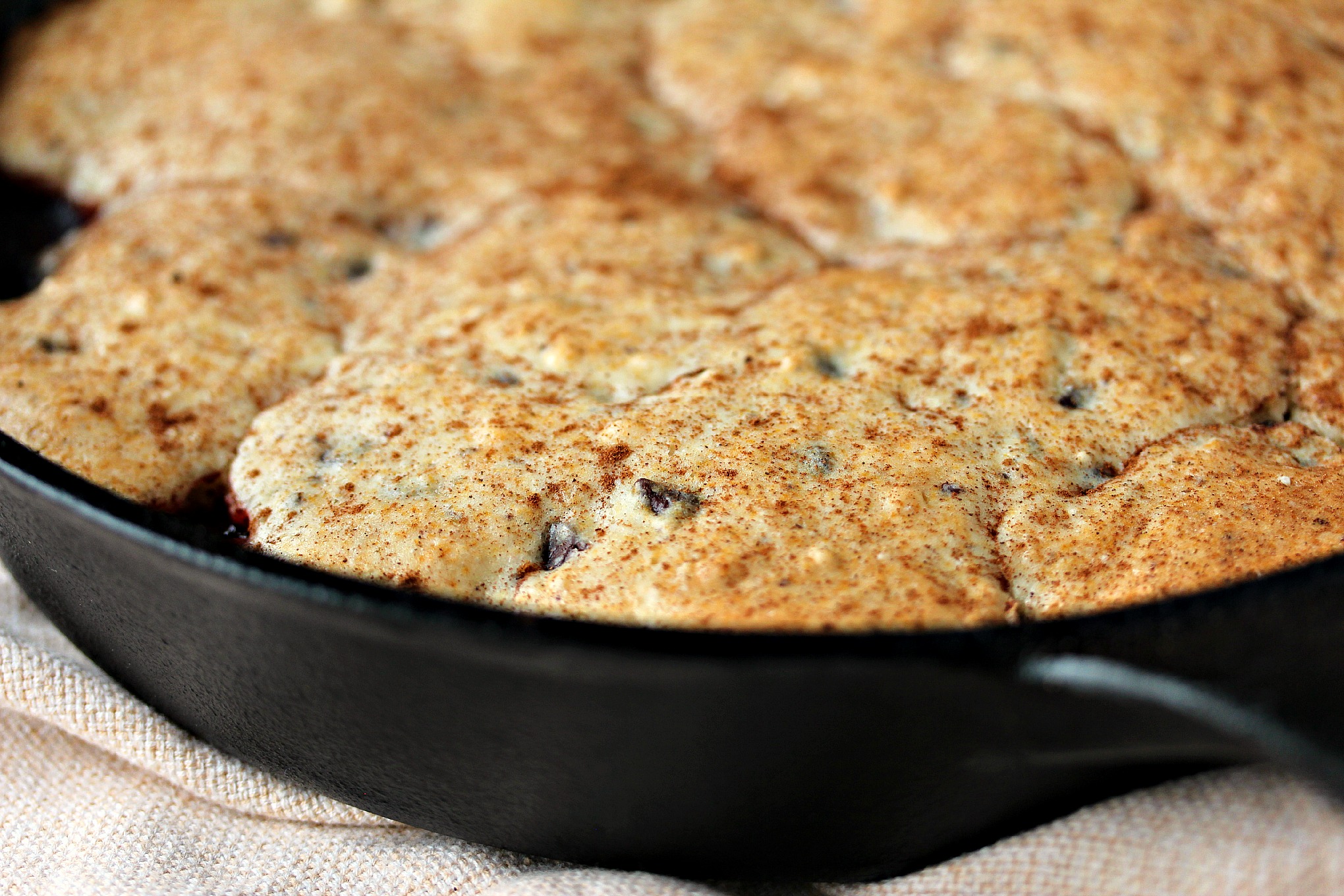 Close up image of cherry cobbler with a chocolate chip muffin topping baked in a cast iron pan and resting on a beige napkin.