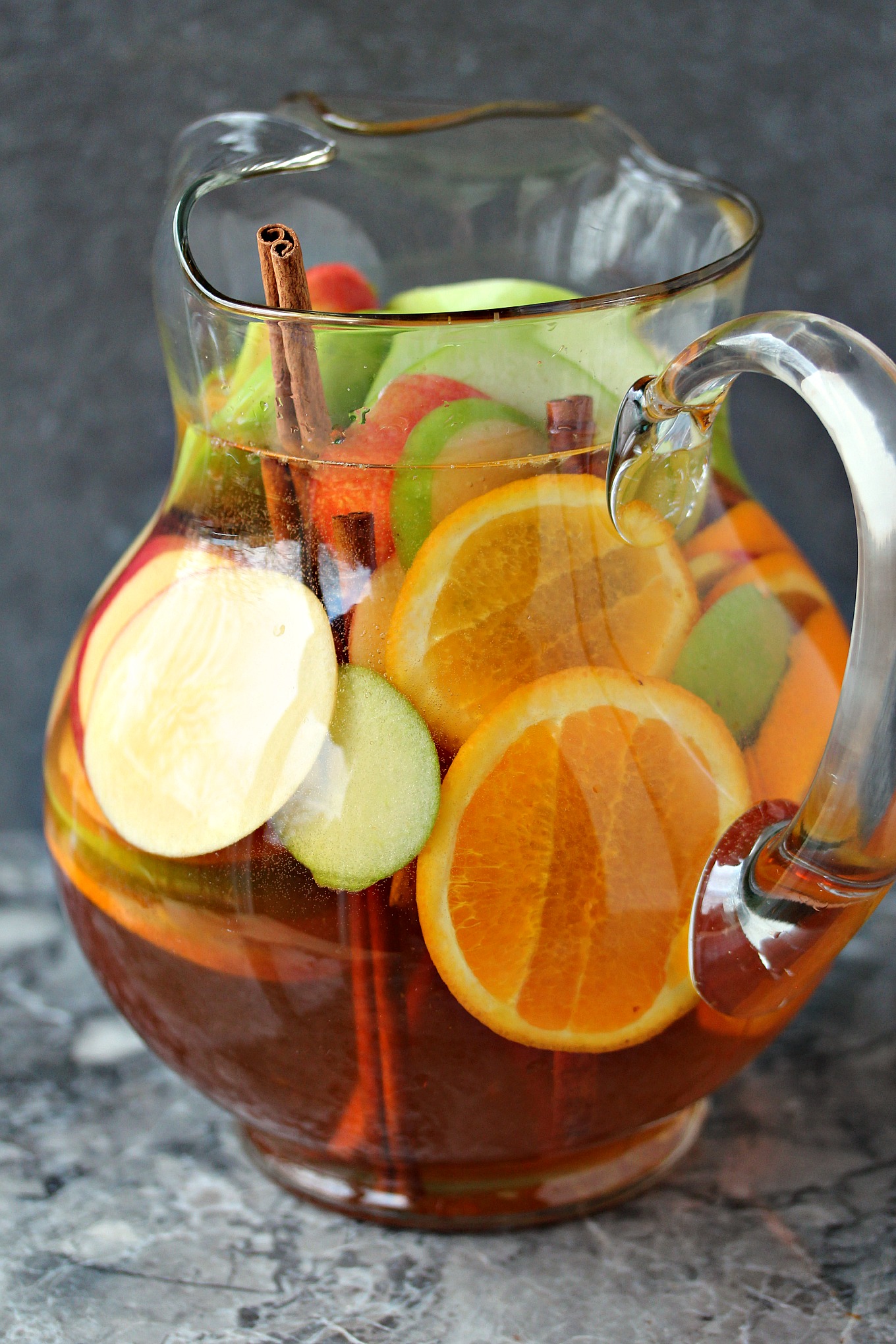 Apple sangria served in a clear glass pitcher with apple slices, orange slices and cinnamon sticks inside. Pitcher is on a grey marble counter.