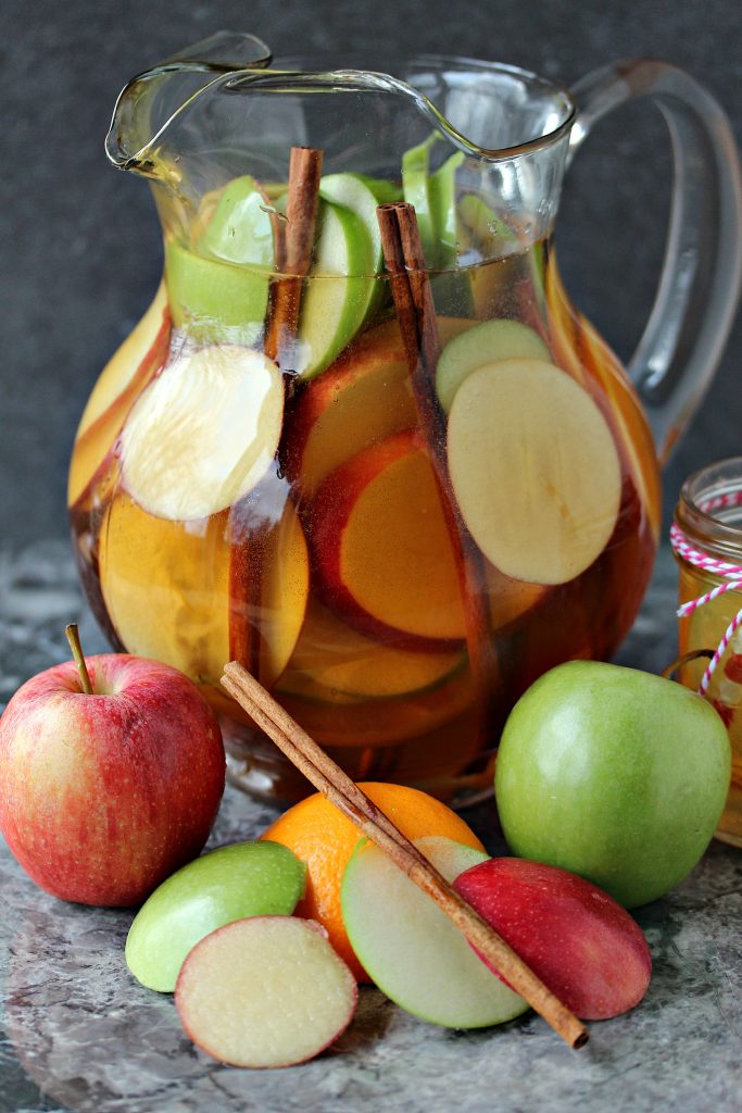 Apple sangria served in a clear glass pitcher with apple slices, orange slices and cinnamon sticks inside. Pitcher is on a grey marble counter with apples, oranges and a spatula on it.