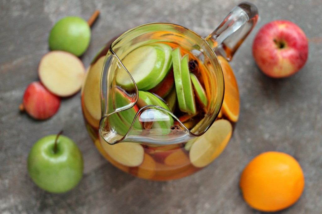 Overhead image of apple sangria in a clear glass pitcher on a neutral coloured countertop with apples, oranges and slices of apples and oranges on it. 