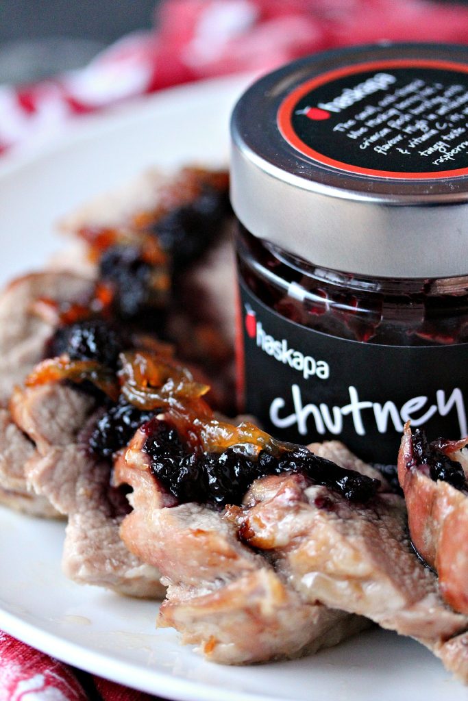 Pork Tenderloin cut int pieces with Haskapa Chutney and caramelized onions over top on a white plate. A jar of Haskapa chutney is in the middle of the plate. 