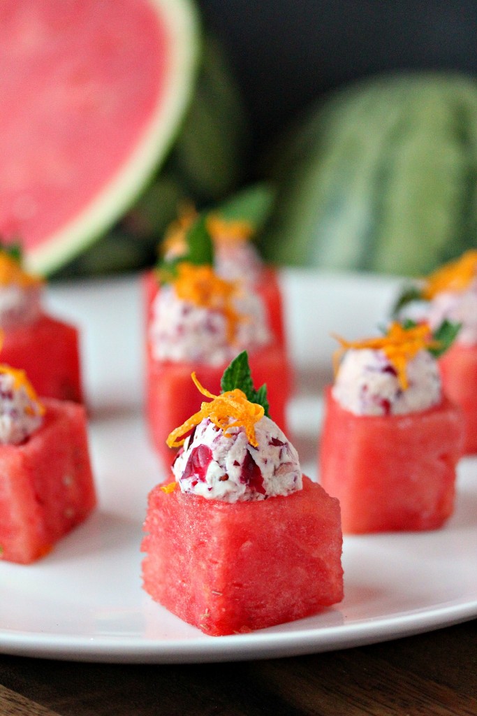 Watermelon Cups with Cranberry Mascarpone scooped inside of them and garnished with lemon zest, all served on a white plate with cut watermelon in the background. 