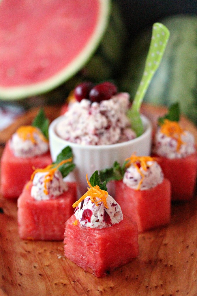 Watermelon Cups with Cranberry Mascarpone scooped inside of them and garnished with lemon zest, all served on a white plate with cut watermelon in the background. Cranberry Mascarpone is also in a small white serving bowl.