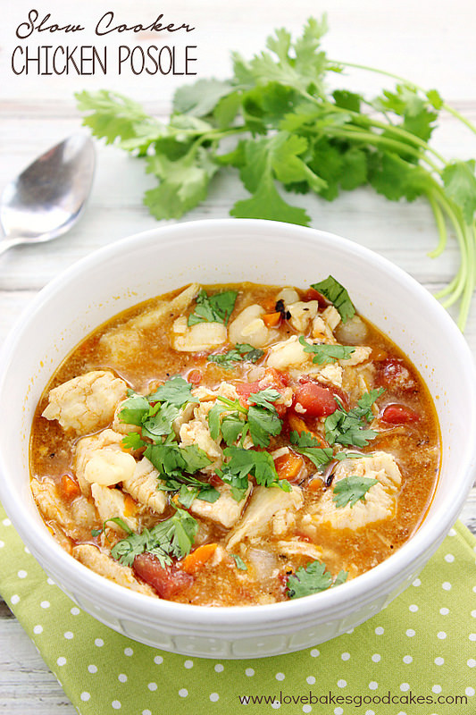 Slow Cooker Chicken Posole – Love Bakes Good Cakes photo image of posole in a white bowl on a green and white polka dot napkin with fresh herbs and a spoon on a white surface. 