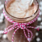A mason jar filled with Nutella hot chocolate, the rim of the jar is tied with twine and there is a silver spoon inside the jar. The hot chocolate is on a neutral counter with tiny marshmallows scattered around.