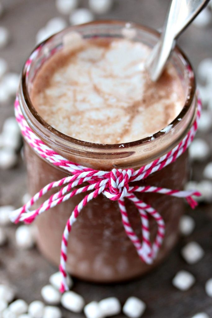 A mason jar filled with Nutella hot chocolate, the rim of the jar is tied with twine and there is a silver spoon inside the jar. The hot chocolate is on a neutral counter with tiny marshmallows scattered around.