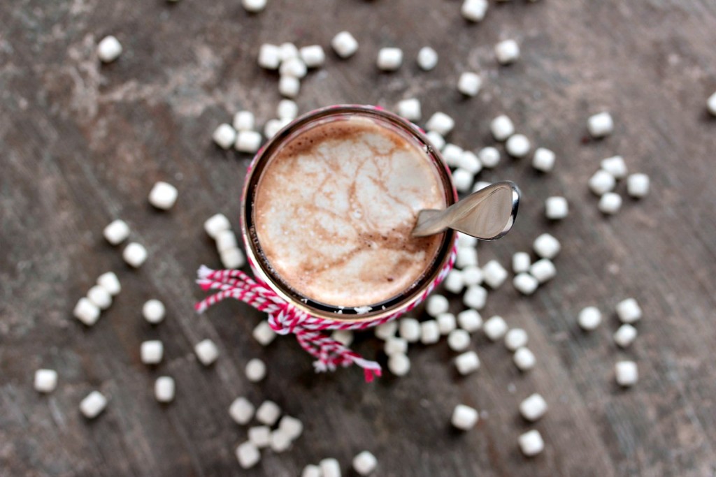 An overhead image of a mason jar filled with Nutella hot chocolate, the rim of the jar is tied with twine, there is a spoon inside the jar, and there are mini marshmallows scattered on the counter. 