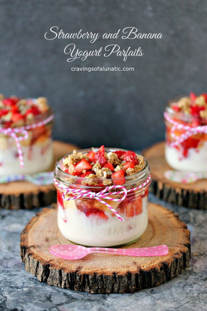 Strawberry and Banana Yogurt Parfaits served in mason jars on wood trivets with colourful spoons resting on them.