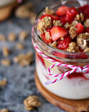 Strawberry and Banana Yogurt Parfaits served in a mason jar on a wood trivet on a grey counter with granola scattered around it.