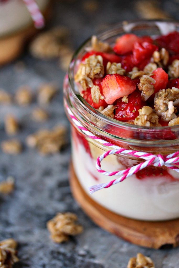Strawberry and Banana Yogurt Parfaits served in a mason jar on a wood trivet on a grey counter with granola scattered around it. 