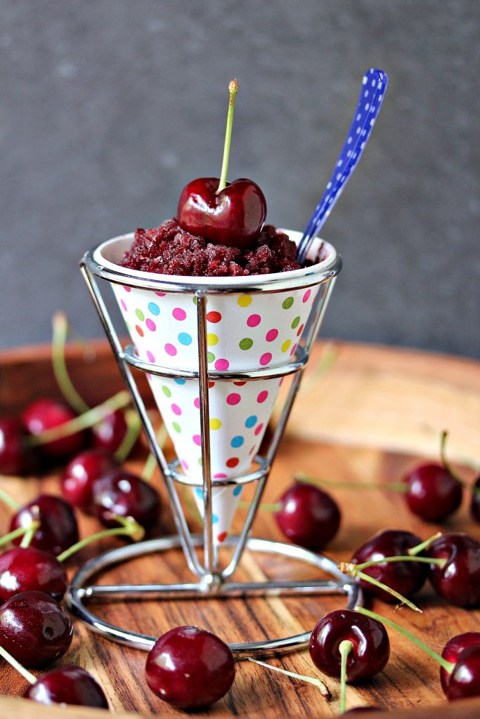 Black cherry granita is served in a white and polka dot paper cone in a metal cone holder on a wood tray with a bottle of Sparkling Ice in black cherry nearby and fresh cherries scattered about. Fresh cherry is on top of the black cherry granita that is in the paper cone. 
