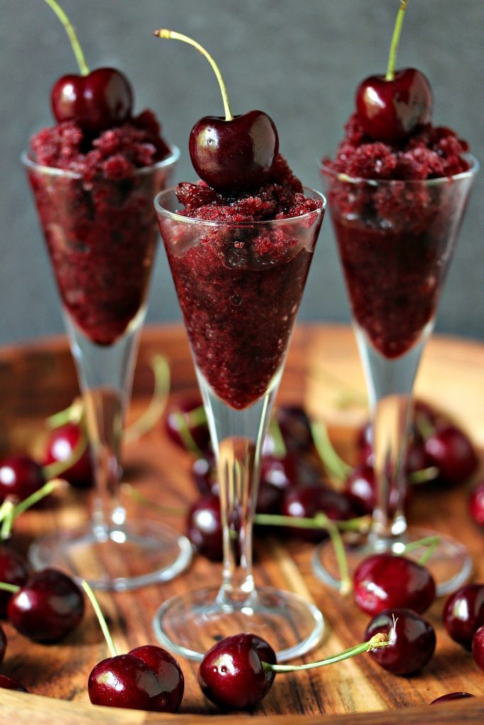 Black cherry granita served in tiny wine glasses with a cherry on top of each granita, cherries are scattered on the wood tray that the glasses are being served on. 
