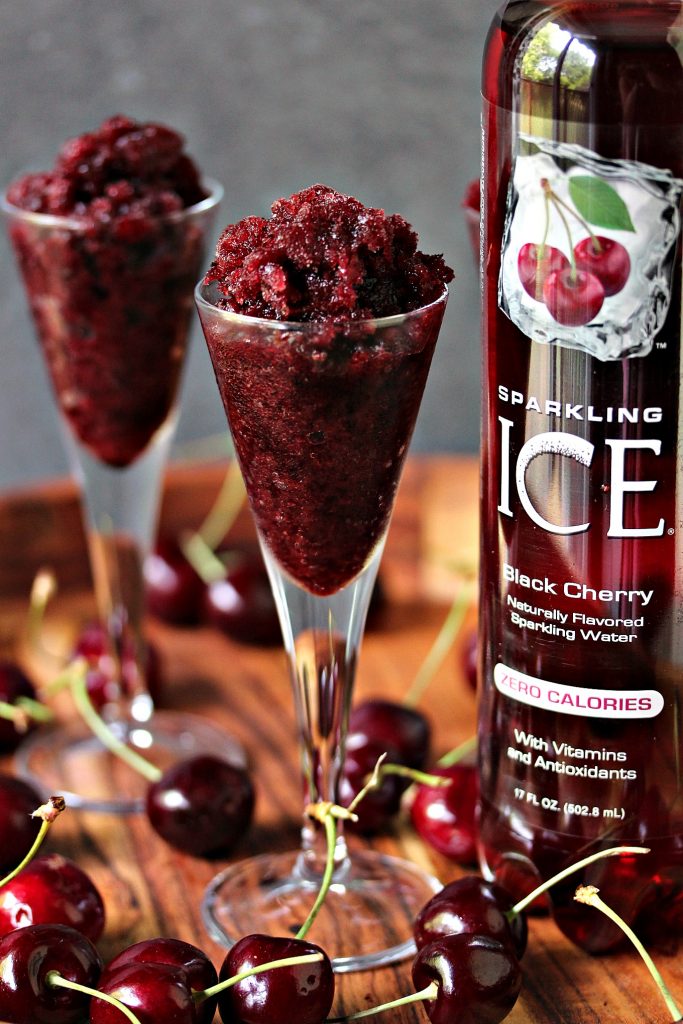 Black cherry granita served in tiny wine glasses with a cherry on top of each granita, cherries are scattered on the wood tray that the glasses are being served on. Bottle of black cherry sparkling ice is on the right side of the photo. 
