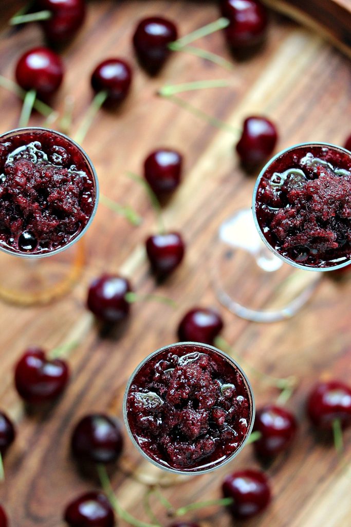 Overhead image of black cherry granita served in tiny wine glasses on a wood board with fresh cherries scattered around the tray.