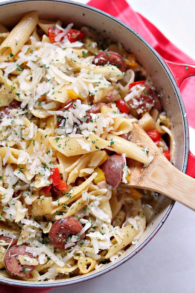 One Pot Pasta with Sausage, Peppers, and Apples in a red Dutch oven with a wooden spoon in it