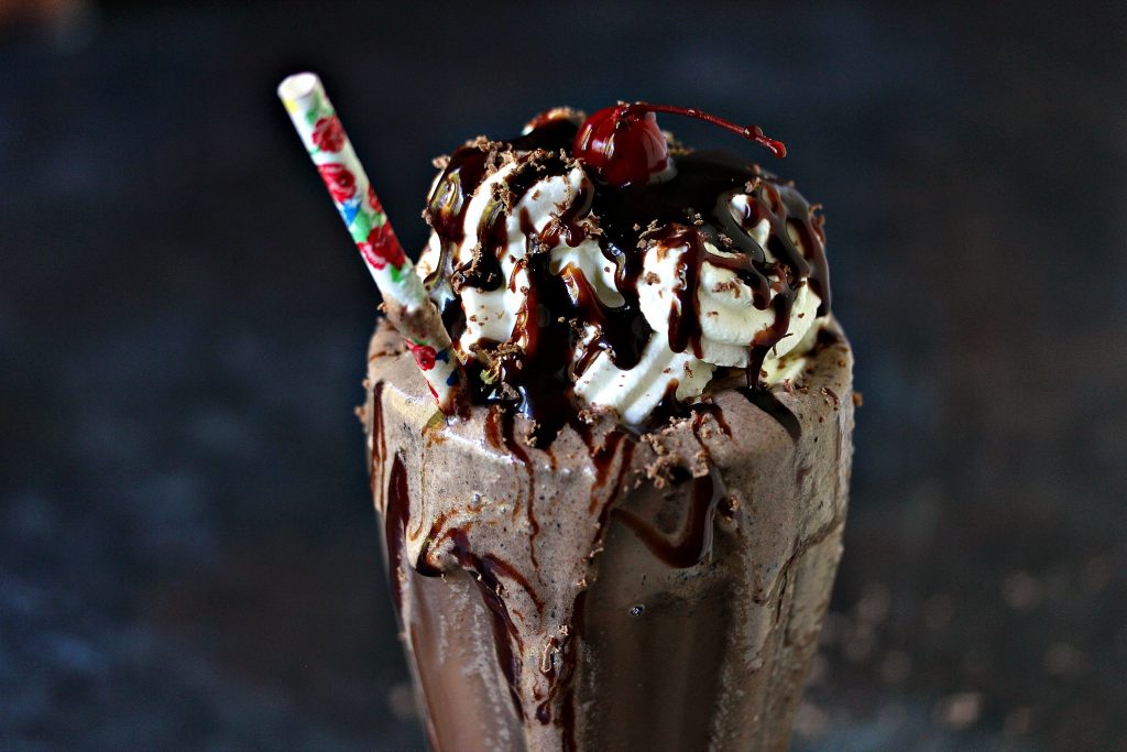  Caramel Macchiato Iced Coffee Chocolate Milkshakes in a milkshake glass with whipped cream, chocolate sauce and a cherry on top. A striped straw is inside the drink and there is a drip of chocolate shake on the front of the glass.