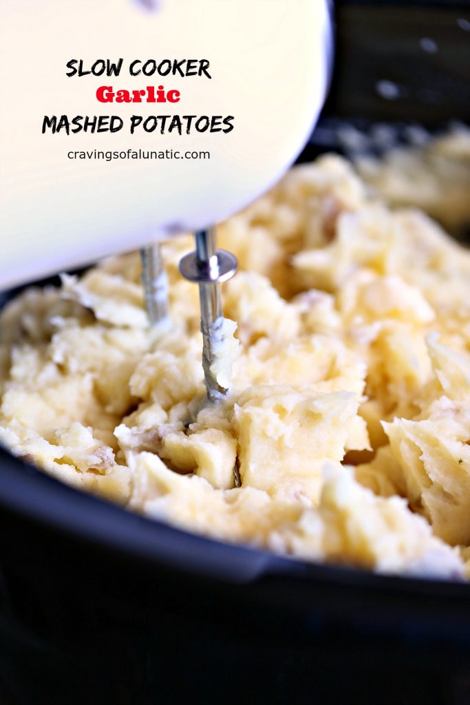 Slow Cooker Garlic Mashed Potatoes in a dark crock pot insert with hand mixer being used to blend the butter and milk into the potatoes. 