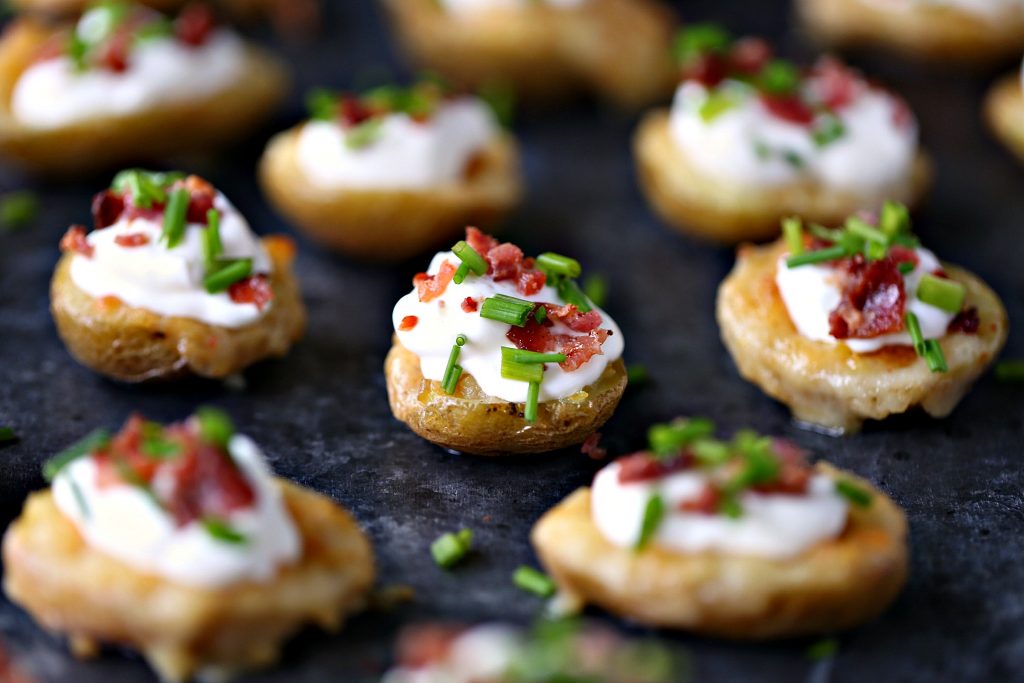 Mini Potato Skins topped with sour cream, chopped bacon and chives on a dark counter.
