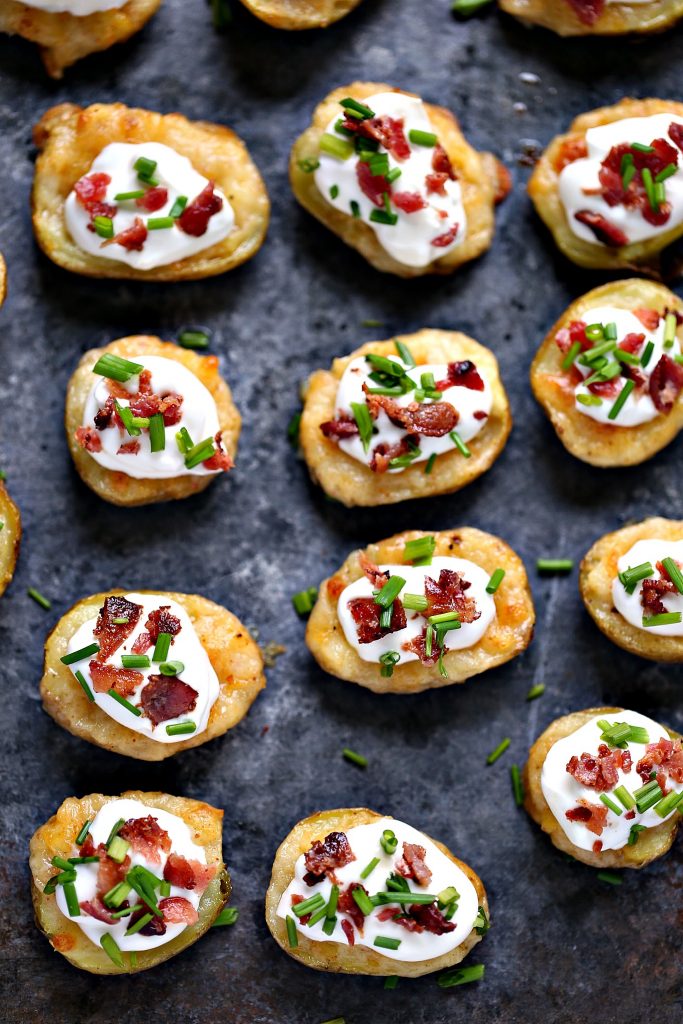 Mini potato skins topped with sour cream, chopped bacon and chives on a dark counter. 