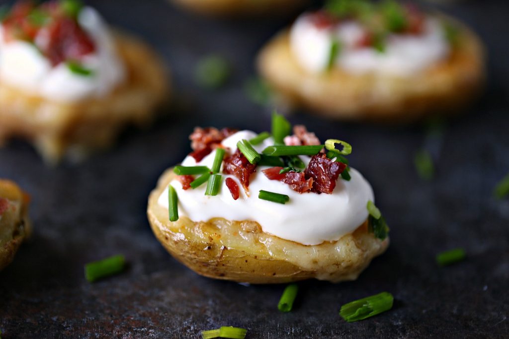 Mini potato skins topped with sour cream, chopped bacon and fresh chives on a dark counter.