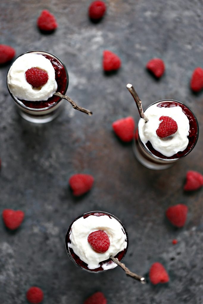 Layered desserts in overhead image, each topped with a fresh raspberry, more raspberries are scattered on a dark surface. 